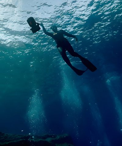 Das Bild zeigt zwei DiverTug Sport Scooter, die auf einem Felsen am Wasser liegen. Im Hintergrund sitzen zwei Taucher am Ufer und beobachten die Szene.